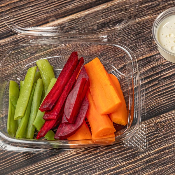 A clear plastic container filled with vibrant green beans, red beets, and orange carrots, with a side of dipping sauce.