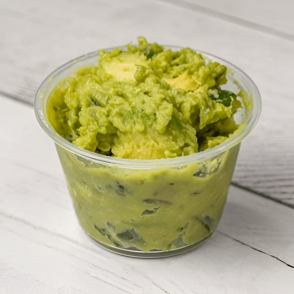 A clear plastic cup filled with chunky green guacamole, featuring visible avocado pieces, on a white wooden surface.